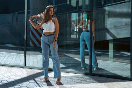 Trendy young woman in white t-shirt in vintage jeans in white shoes walks on city. Attractive girl in casual clothes is walking on the street. american style.の写真素材