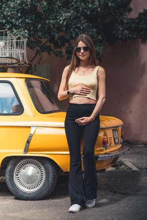 Happy pregnant young girl with a smile in a fashionable knit tank top and dark jeans with glasses is standing near a yellow retro car. Female summer style outfitの写真素材