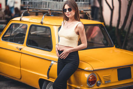 Happy pregnant young girl with a smile in a fashionable knit tank top and dark jeans with glasses is standing near a yellow retro car. Female summer style outfitの写真素材