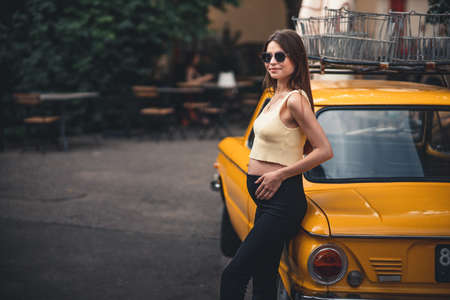 Happy pregnant young girl with a smile in a fashionable knit tank top and dark jeans with glasses is standing near a yellow retro car. Female summer style outfitの写真素材