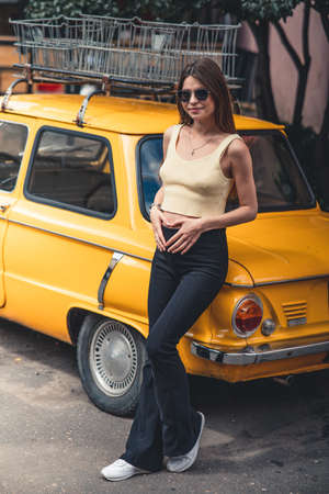 Happy pregnant young girl with a smile in a fashionable knit tank top and dark jeans with glasses is standing near a yellow retro car. Female summer style outfitの写真素材