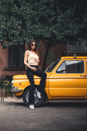 Happy pregnant young girl with a smile in a fashionable knit tank top and dark jeans with glasses is standing near a yellow retro car. Female summer style outfitの写真素材