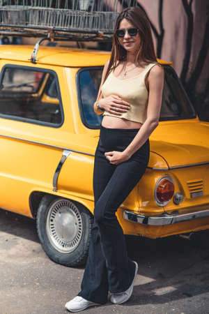 Happy pregnant young girl with a smile in a fashionable knit tank top and dark jeans with glasses is standing near a yellow retro car. Female summer style outfitの写真素材