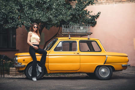 Happy pregnant young girl with a smile in a fashionable knit tank top and dark jeans with glasses is standing near a yellow retro car. Female summer style outfitの写真素材