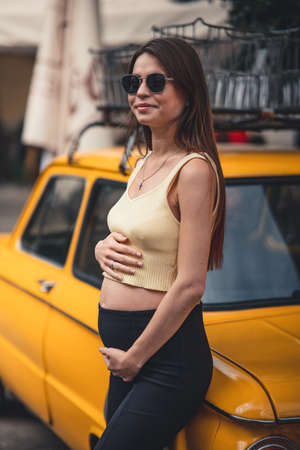 Happy pregnant young girl with a smile in a fashionable knit tank top and dark jeans with glasses is standing near a yellow retro car. Female summer style outfitの写真素材