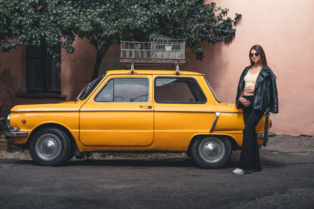 Happy pregnant young girl with a smile in a fashionable knit tank top and dark jeans with glasses is standing near a yellow retro car. Female summer style outfitの写真素材