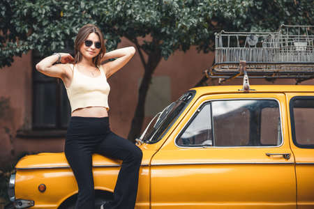 Happy pregnant young girl with a smile in a fashionable knit tank top and dark jeans with glasses is standing near a yellow retro car. Female summer style outfitの写真素材
