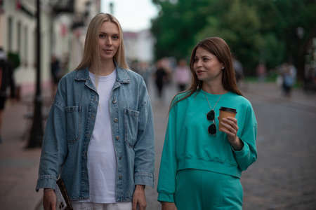 Two cool fashionable pregnant girlfriends in fashionable clothes are walking down the street and talking. Friends share secrets and discuss the birth of children.の写真素材