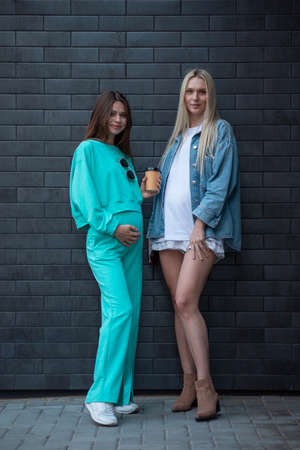 Two cool fashionable pregnant girlfriends in fashionable clothes are walking down the street and talking. Friends share secrets and discuss the birth of children.の写真素材