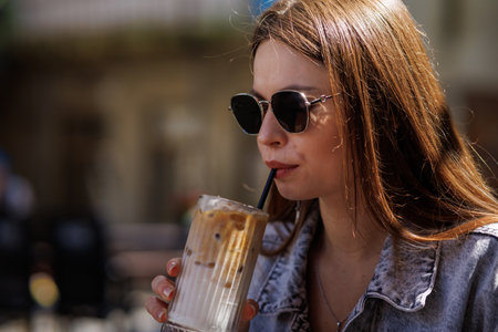 Pretty young hipster woman in stylish glamorous black glasses in trendy blue denim jacket with coffee. American girl fashion model posing in the city.の写真素材