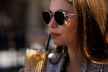 Pretty young hipster woman in stylish glamorous black glasses in trendy blue denim jacket with coffee. American girl fashion model posing in the city.の写真素材