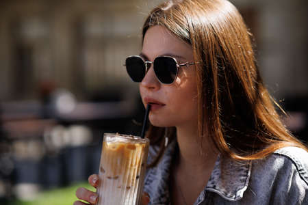Pretty young hipster woman in stylish glamorous black glasses in trendy blue denim jacket with coffee. American girl fashion model posing in the city.の写真素材