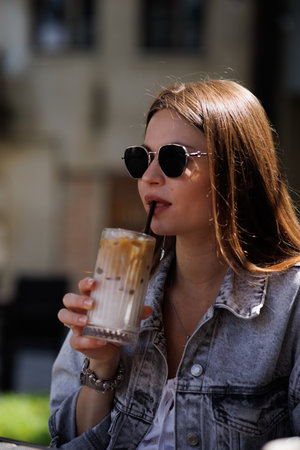 Pretty young hipster woman in stylish glamorous black glasses in trendy blue denim jacket with coffee. American girl fashion model posing in the city.の写真素材