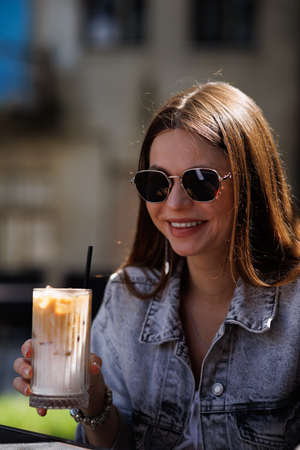 Pretty young hipster woman in stylish glamorous black glasses in trendy blue denim jacket with coffee. American girl fashion model posing in the city.の写真素材