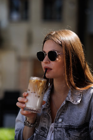 Pretty young hipster woman in stylish glamorous black glasses in trendy blue denim jacket with coffee. American girl fashion model posing in the city.の写真素材