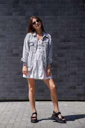 Fashionable women's portrait of a beautiful vogue girl pregnant model with fashion black eyewear in a stylish white short dress near a black brick wallの写真素材