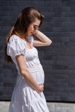 Fashionable women's portrait of a beautiful vogue girl pregnant model with fashion black eyewear in a stylish white short dress near a black brick wallの写真素材