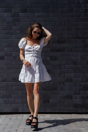 Fashionable women's portrait of a beautiful vogue girl pregnant model with fashion black eyewear in a stylish white short dress near a black brick wallの写真素材