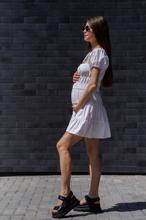 Fashionable women's portrait of a beautiful vogue girl pregnant model with fashion black eyewear in a stylish white short dress near a black brick wallの写真素材