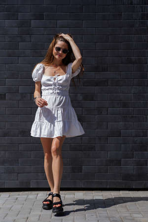 Fashionable women's portrait of a beautiful vogue girl pregnant model with fashion black eyewear in a stylish white short dress near a black brick wallの写真素材