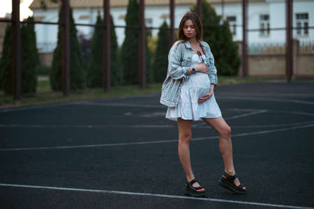 Beautiful young pregnant girl in a white dress and a denim jacket on a basketball court. The woman gently holds her hands on her stomach. The concept of pregnancy and sports.の写真素材