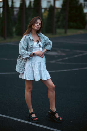 Beautiful young pregnant girl in a white dress and a denim jacket on a basketball court. The woman gently holds her hands on her stomach. The concept of pregnancy and sports.の写真素材