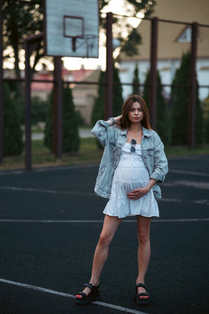 Beautiful young pregnant girl in a white dress and a denim jacket on a basketball court. The woman gently holds her hands on her stomach. The concept of pregnancy and sports.の写真素材