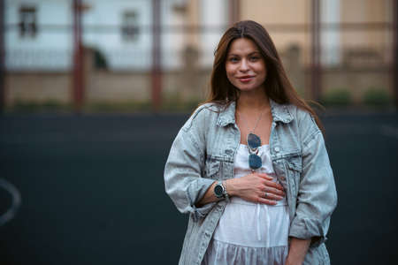 Beautiful young pregnant girl in a white dress and a denim jacket on a basketball court. The woman gently holds her hands on her stomach. The concept of pregnancy and sports.の写真素材
