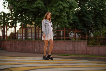 Beautiful happy pregnant woman in a vintage fashion jeans jacket and white dress walking in the city.の写真素材