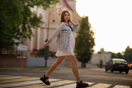 Beautiful happy pregnant woman in a vintage fashion jeans jacket and white dress walking in the city.の写真素材