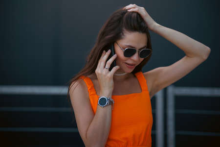 Serious business woman with black sunglasses in fashion orange dress talking on the phone and walking in city.の写真素材