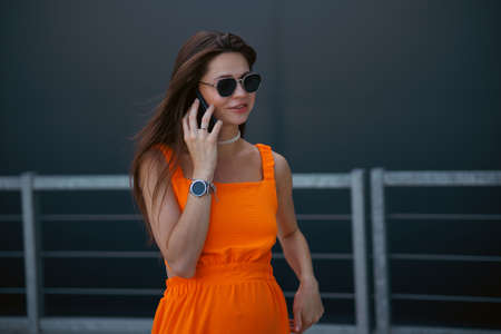 Serious business woman with black sunglasses in fashion orange dress talking on the phone and walking in city.の写真素材