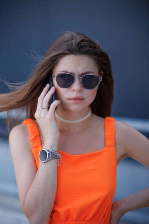 Serious business woman with black sunglasses in fashion orange dress talking on the phone and walking in city.の写真素材