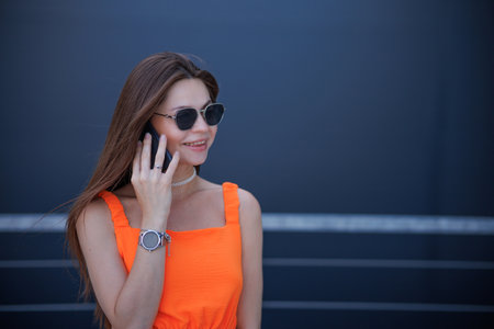 Serious business woman with black sunglasses in fashion orange dress talking on the phone and walking in city.の写真素材