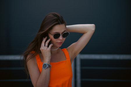Serious business woman with black sunglasses in fashion orange dress talking on the phone and walking in city.の写真素材