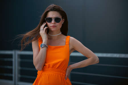 Serious business woman with black sunglasses in fashion orange dress talking on the phone and walking in city.の写真素材
