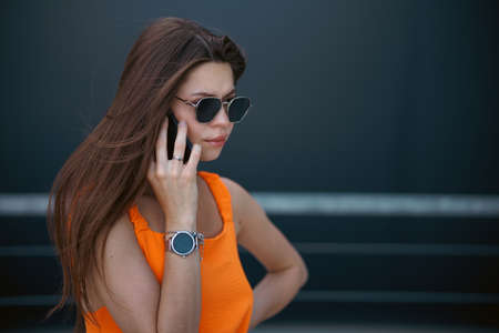 Serious business woman with black sunglasses in fashion orange dress talking on the phone and walking in city.の写真素材