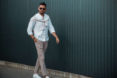 Fashion model handsome young man with trendy hairstyle in sunglasses in white shirt in light jeans posing near vintage wall on street on sunny day. Attractive american guy on walk.の写真素材