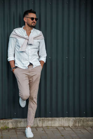 Fashion model handsome young man with trendy hairstyle in sunglasses in white shirt in light jeans posing near vintage wall on street on sunny day. Attractive american guy on walk.の写真素材