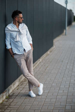 Fashion model handsome young man with trendy hairstyle in sunglasses in white shirt in light jeans posing near vintage wall on street on sunny day. Attractive american guy on walk.の写真素材