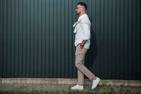 Fashion model handsome young man with trendy hairstyle in sunglasses in white shirt in light jeans posing near vintage wall on street on sunny day. Attractive american guy on walk.の写真素材