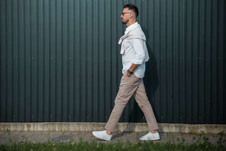 Fashion model handsome young man with trendy hairstyle in sunglasses in white shirt in light jeans posing near vintage wall on street on sunny day. Attractive american guy on walk.の写真素材