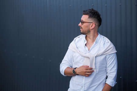 Fashion model handsome young man with trendy hairstyle in sunglasses in white shirt in light jeans posing near vintage wall on street on sunny day. Attractive american guy on walk.の写真素材