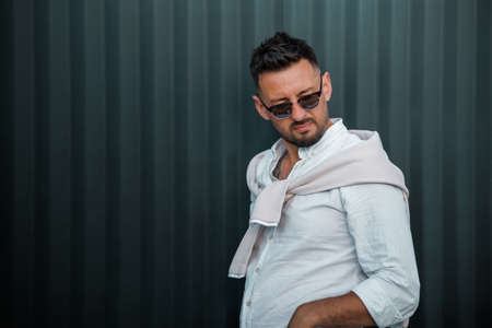Fashion model handsome young man with trendy hairstyle in sunglasses in white shirt in light jeans posing near vintage wall on street on sunny day. Attractive american guy on walk.の写真素材