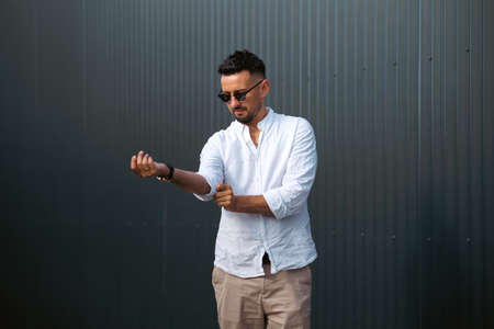 Fashion model handsome young man with trendy hairstyle in sunglasses in white shirt in light jeans posing near vintage wall on street on sunny day. Attractive american guy on walk.の写真素材