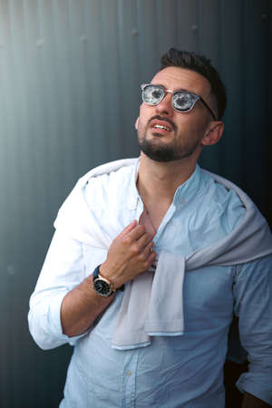 Fashion model handsome young man with trendy hairstyle in sunglasses in white shirt in light jeans posing near vintage wall on street on sunny day. Attractive american guy on walk.の写真素材
