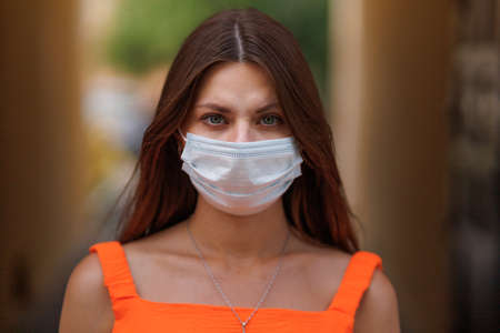 Girl, young woman in protective sterile medical mask on her face looking at camera outdoors close up. corona virus pandemic. Beautiful brunette haired girl with medical mask.の写真素材