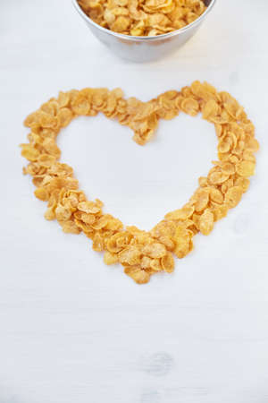 Cornflakes in a metal bowl on a painted white wooden background. The symbol of the heart is laid out of cornflakes.の写真素材