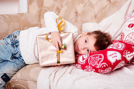 Cute little boy is lying on the sofa with a gift in his hands. A guy with a stylish Iroquois haircut and in trendy modern clothes. The boy is holding a large gift box with a bowの写真素材