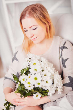 Beautiful young woman sits tilted head and holds a bouquet of flowers. Portrait of a pretty girl in a light knitted sweater with a pattern of stars. Woman with blond hair keep holding white daisiesの写真素材
